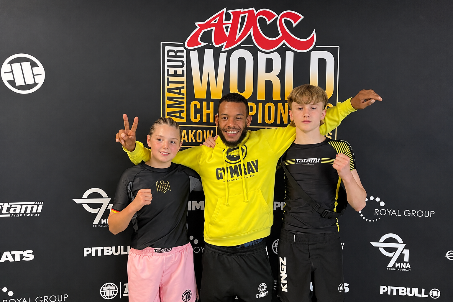 Lenny Arend und Joleen Bugge von GYMRAY Bamberg mit Coach Ray vor der Fotowand der ADCC Amateur World Championship 2026 in Krakau