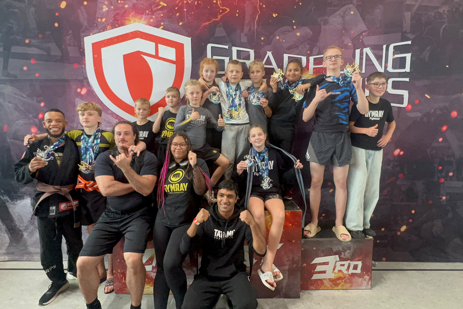 Das Team von GYMRAY Bamberg mit Athleten, Coaches und Medaillen auf dem Siegerpodest vor der Grappling-Industries-Wand in Nürnberg