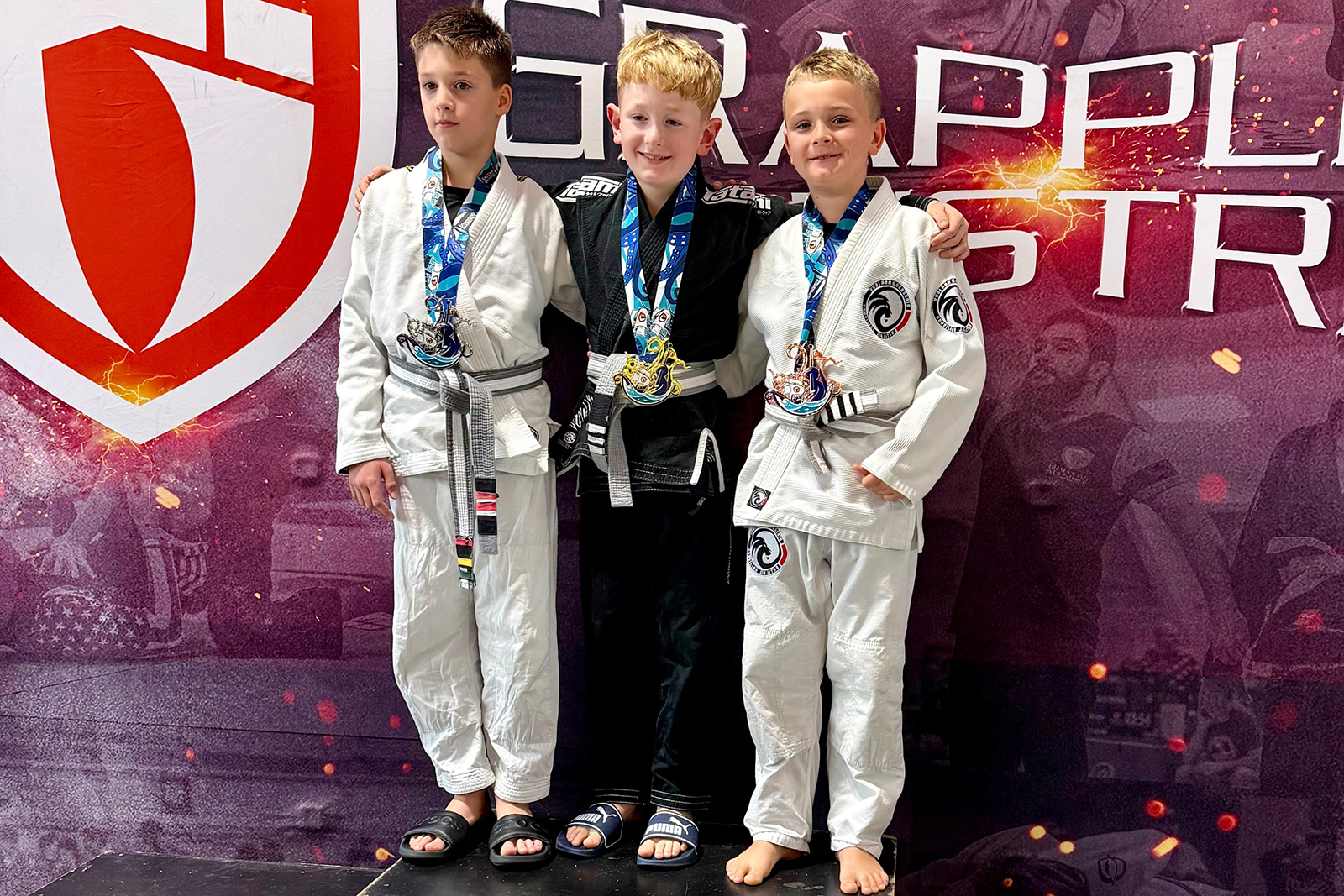 Max Ecker vom GYMRAY Kampfsport Bamberg im schwarzen Gi mit Goldmedaille in der Mitte des Siegerpodests bei Grappling Industries Nürnberg, links und rechts zwei Kinder in weißen Gis mit Silber- und Bronzemedaille.